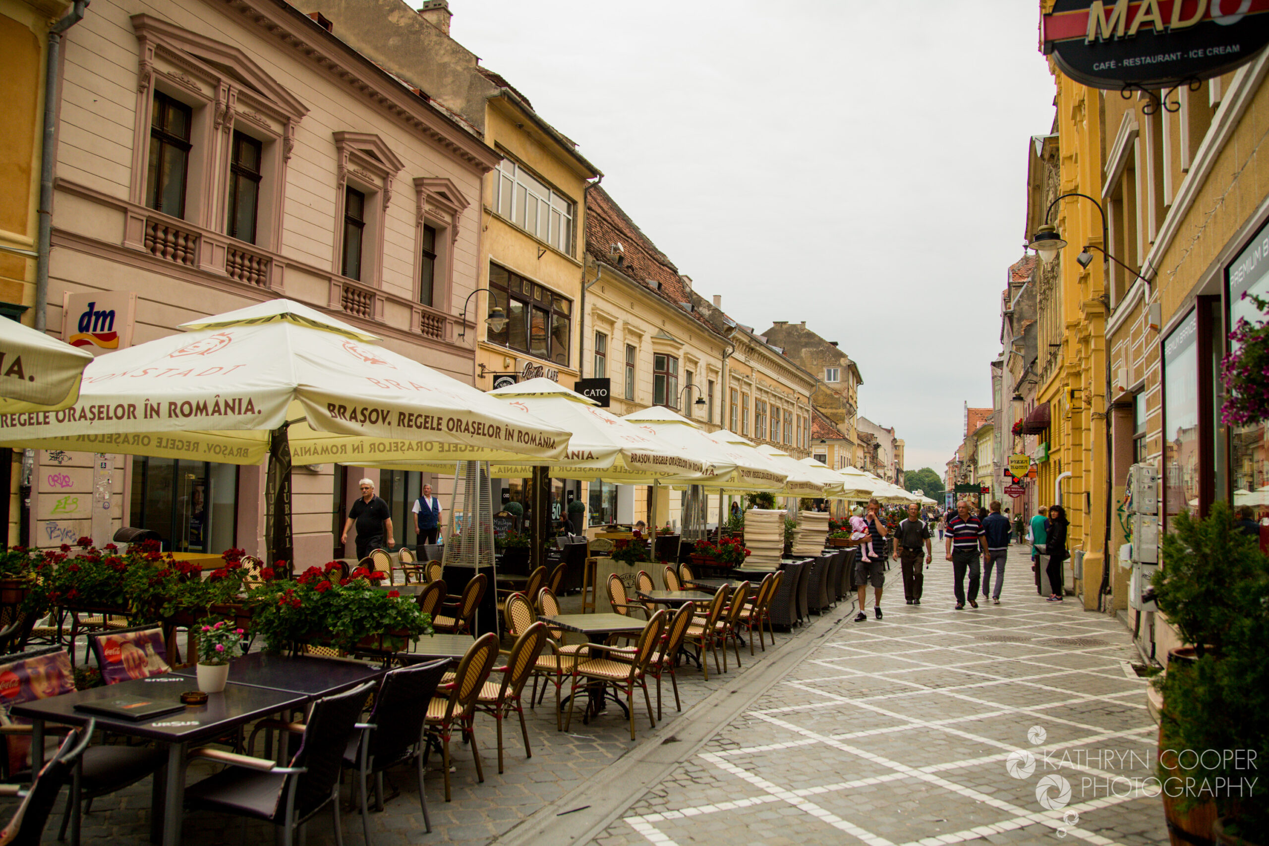 A line of bar terraces with seating and umbrellas sprawl along a cobbled street in old town Brasov.