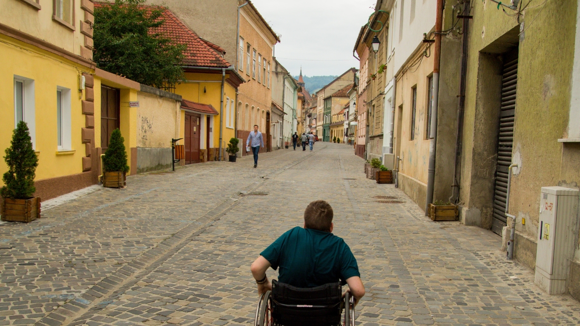 A wheelchair user, with his back to camera, pushes himself through one of the many side streets in Brasov.