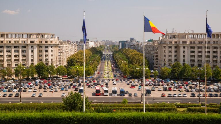 A view from the peoples palace in Bucharest, looking down a long boulevard, with parked cars and flags raised.
