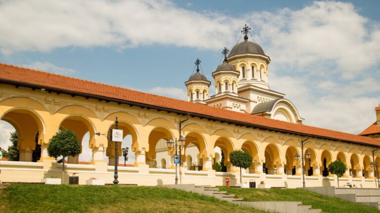 The Coronation Cathedral in Alba Iulia, Romania, seen on a sunny day. The building features cream-colored walls, red-tiled roofs, and black-domed towers with crosses. A row of arches and neatly trimmed trees line the walkway in front, with a blue accessible toilet sign visible near a lamppost.