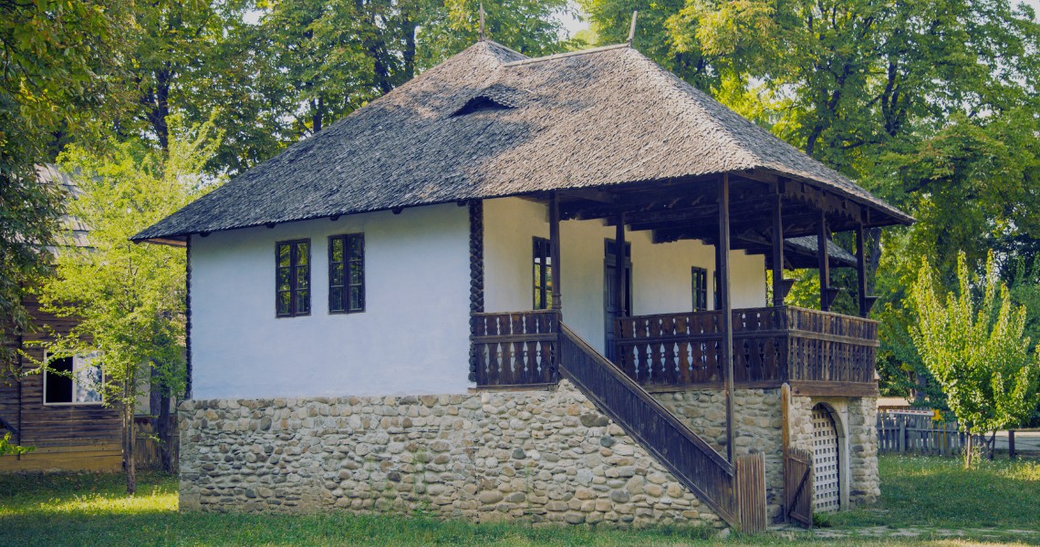 A traditional whitewashed Romanian house with a wraparound wooden porch, carved railings, and a stone staircase, set among trees.