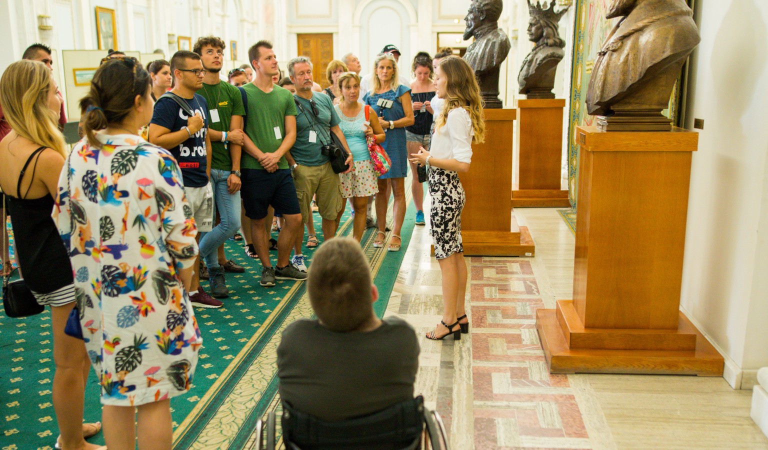 A wheelchair user, seen from behind, joins a tour group inside the Palace of the Parliament in Bucharest, listening to a guide speak in a corridor lined with bronze busts on wooden pedestals. The group stands on a patterned marble and green-carpeted floor, with ornate white walls and framed portraits visible in the background.