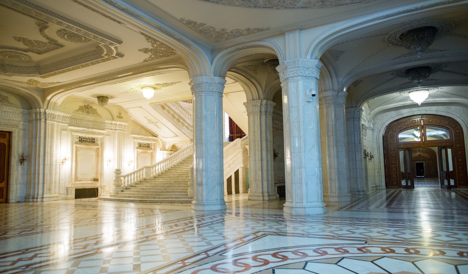 A grand marble interior hall of the Palace of the Parliament in Bucharest, with white marble columns, an ornately decorated gilded ceiling, and a wide curved staircase with balustrades in the background. The polished floor features an intricate inlaid marble pattern, and chandeliers hang from the corridor to the right, which leads toward a large wooden door.
