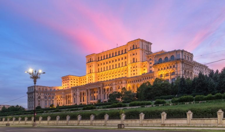 The Palace of the Parliament in Bucharest, Romania, illuminated in golden light at dusk, with a vivid pink and purple sunset sky behind it. The grand neoclassical building features rows of arched windows and columns, set behind manicured lawns, hedges, and a stone perimeter fence with an ornate lamppost in the foreground.